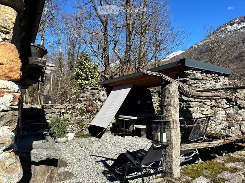 A rustic outdoor seating area with stone walls, a covered seating area, tables, chairs, and a lantern, surrounded by gravel and greenery with a mountain view.