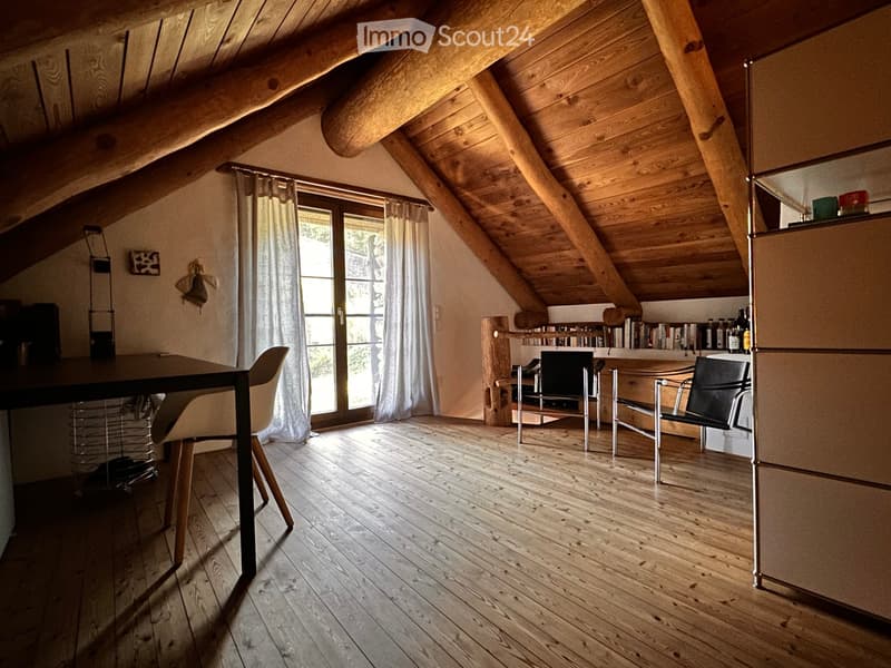 Bright attic room with wooden beams, wooden floor, large window with curtains, and a small kitchen area with a bookshelf and cupboard.
