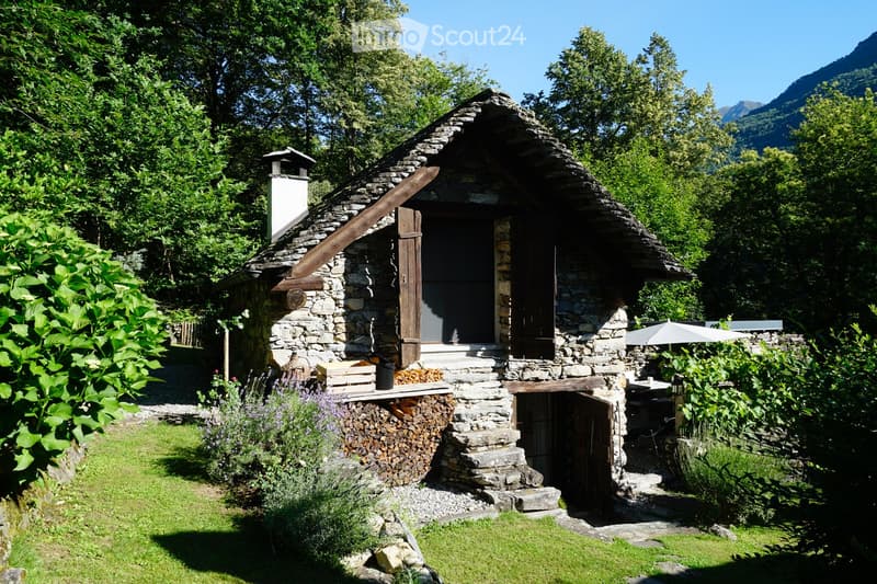 A rustic stone cottage with a steep, slate roof, surrounded by lush greenery and a scenic mountain backdrop. Features include a chimney, wooden shutters, a small porch, and a neatly stacked woodpile.