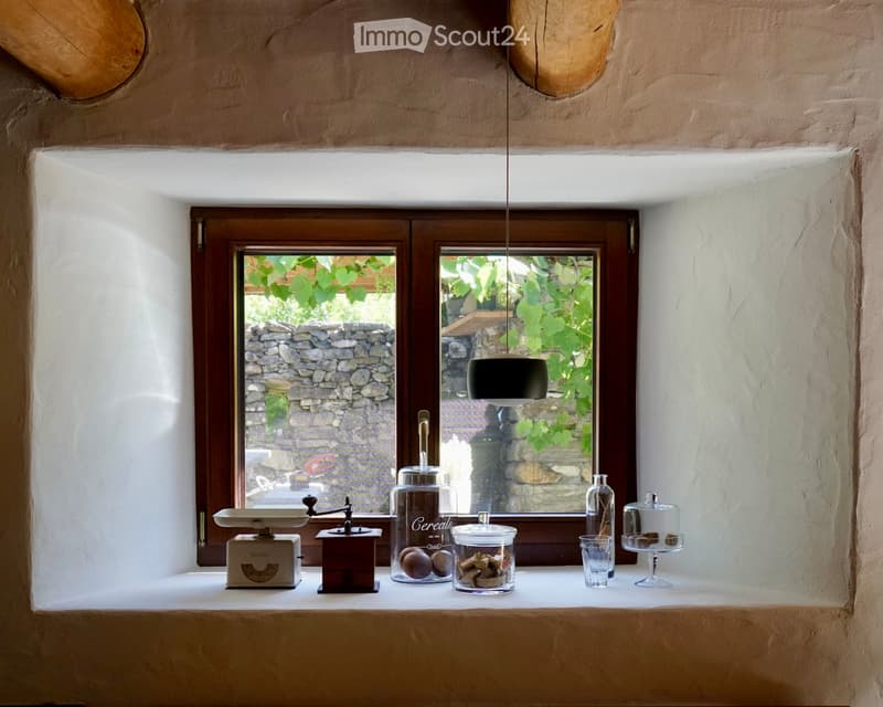 Window sill with a scale, coffee grinder, glass jars, a glass bottle, a cup, and a cake, overlooking a stone wall and greenery.