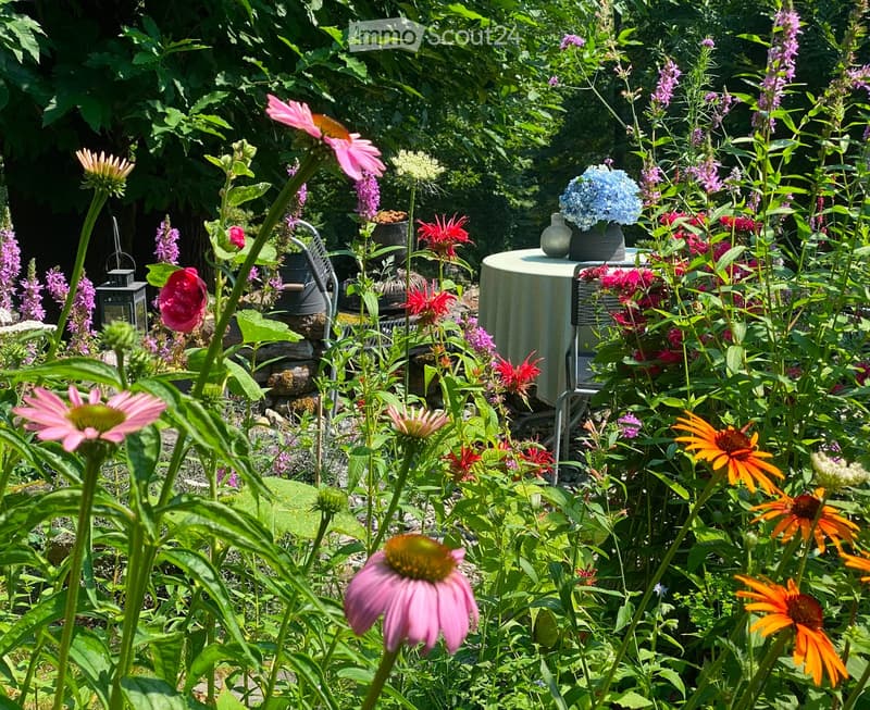 A lush garden filled with colorful flowers, including pink, orange, and purple blooms, surrounded by greenery and a small table with vases.