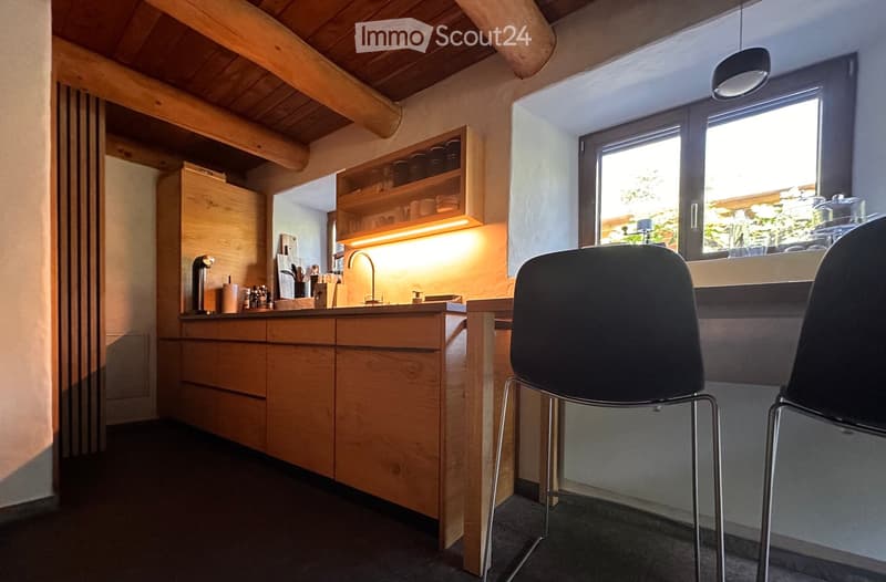 Modern kitchen with wooden cabinets, a sink, and a window overlooking greenery. Two stools are placed near the counter, and the space features built-in storage and a sleek design.