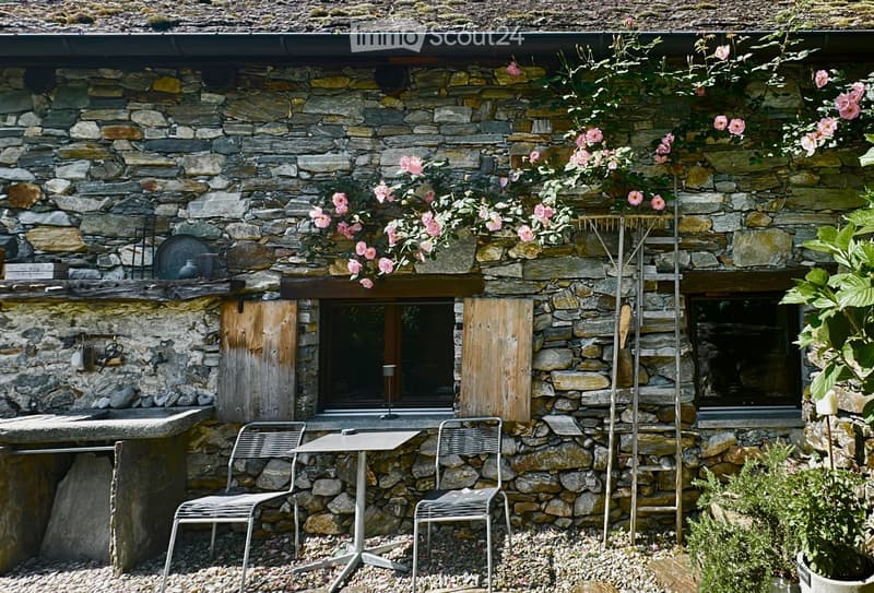 Stone cottage exterior with a small outdoor seating area featuring a metal table and two chairs, surrounded by flowering plants and greenery.