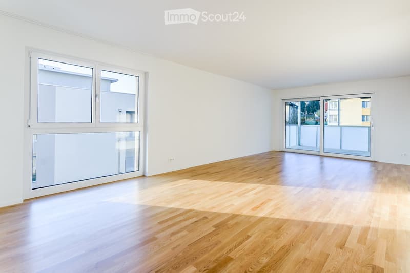 Empty room with light wooden flooring, white walls, large windows, and sliding glass doors leading to a balcony.