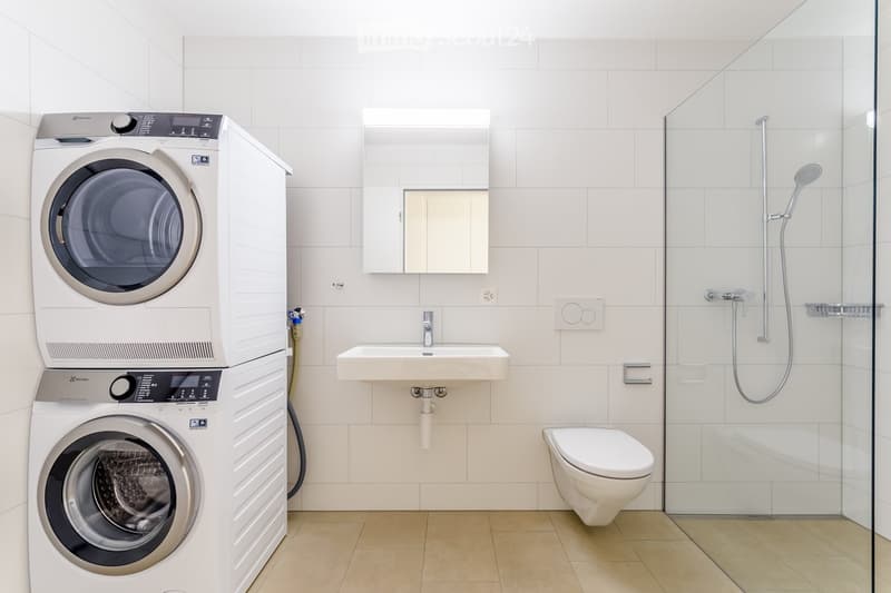 Modern bathroom with white tiles, a stacked washer and dryer unit, a sink with mirror, a toilet, and a glass-enclosed shower with a handheld showerhead.