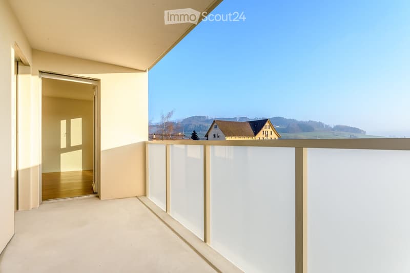 Balcony with white railing, open sliding door leading to a room, scenic countryside view.
