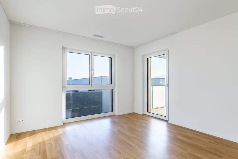 Empty room with white walls, wooden flooring, large windows, and a sliding glass door leading outside.