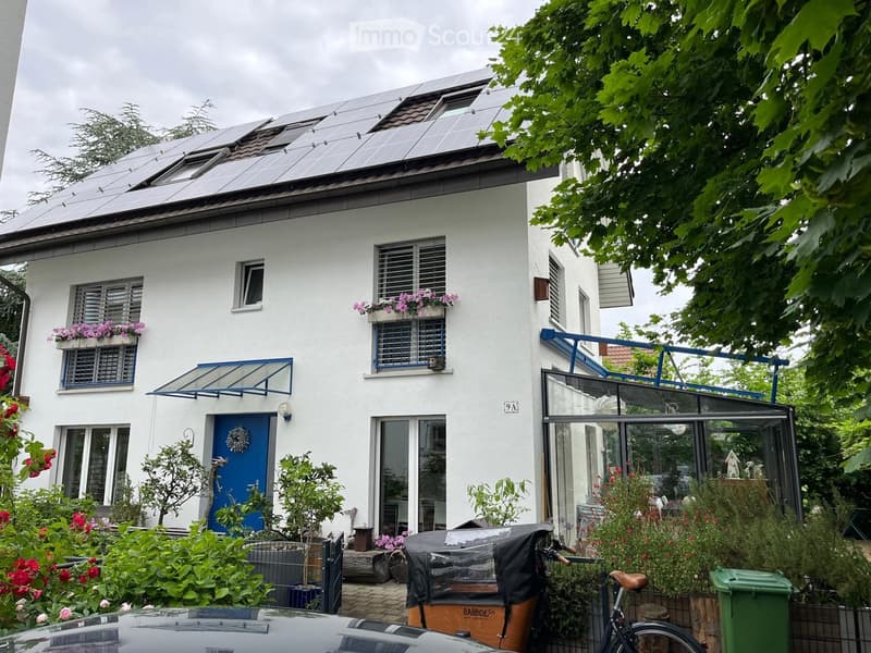 Two-story white house with solar panels on the roof, blue shutters, and flower boxes on the windows. Features a covered entrance, a bicycle, and a small garden area with plants and a trash bin.