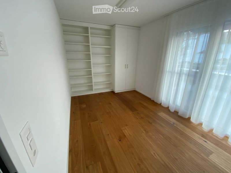 Empty room with wooden flooring, white walls, built-in shelving unit, and large window with white curtains.