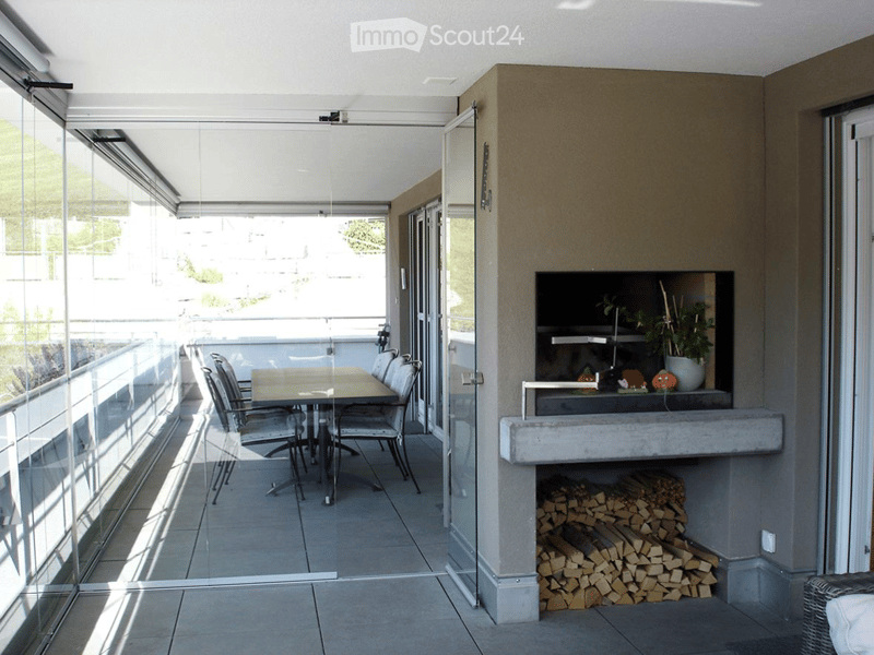 Modern balcony with glass sliding doors, tiled floor, dining table with chairs, fireplace with stacked firewood, and decorative items on mantel.