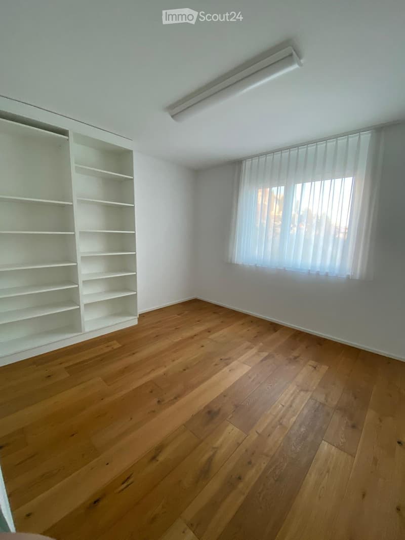 Empty room with wooden flooring, white walls, built-in shelving unit, and a window with sheer curtains.