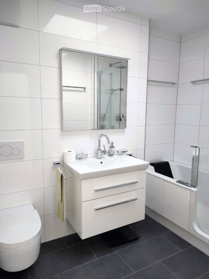 Modern bathroom with white tiles, a white sink with a chrome faucet, a large mirror, a toilet, and a bathtub with a glass shower enclosure.