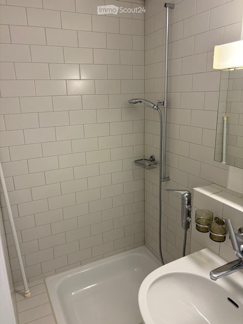 Modern bathroom with light gray tiled walls, a bathtub, a sink with a modern faucet, a showerhead with a handheld hose, and a small shower rack holding two glass cups.