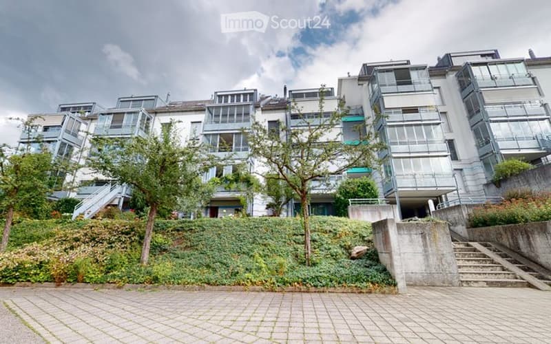 Modern multi-story residential building with large windows and balconies, surrounded by greenery and a paved walkway.