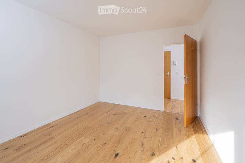 Empty room with light wooden flooring and white walls, featuring an open wooden door leading to another room.