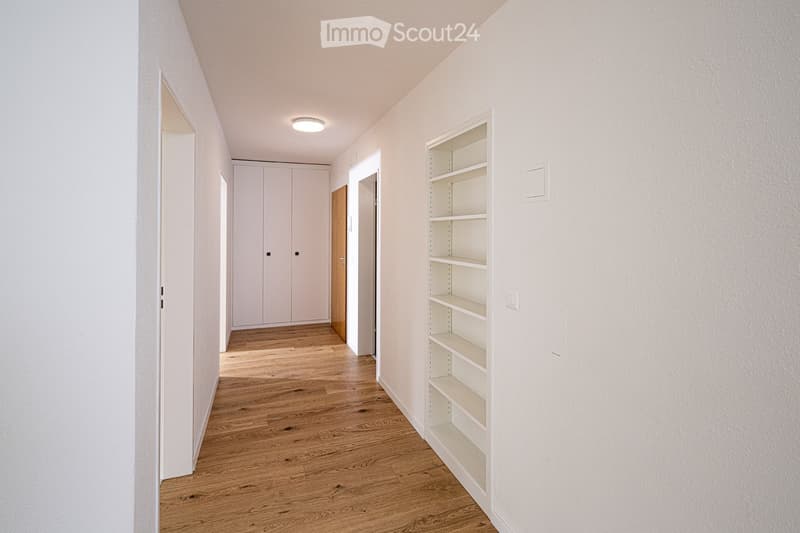A clean, modern hallway with light wooden flooring, white walls, built-in shelving on one side, and a closed cabinet at the end.