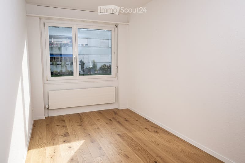 Empty room with wooden flooring, white walls, a large window with a view of a balcony area, and a white radiator below the window.