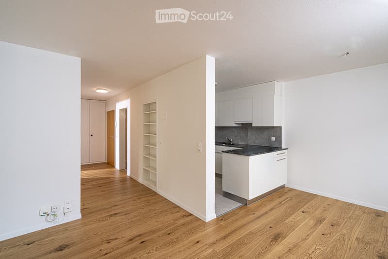 Empty hallway with wooden flooring, white walls, and a small kitchen area with white cabinets and a countertop.