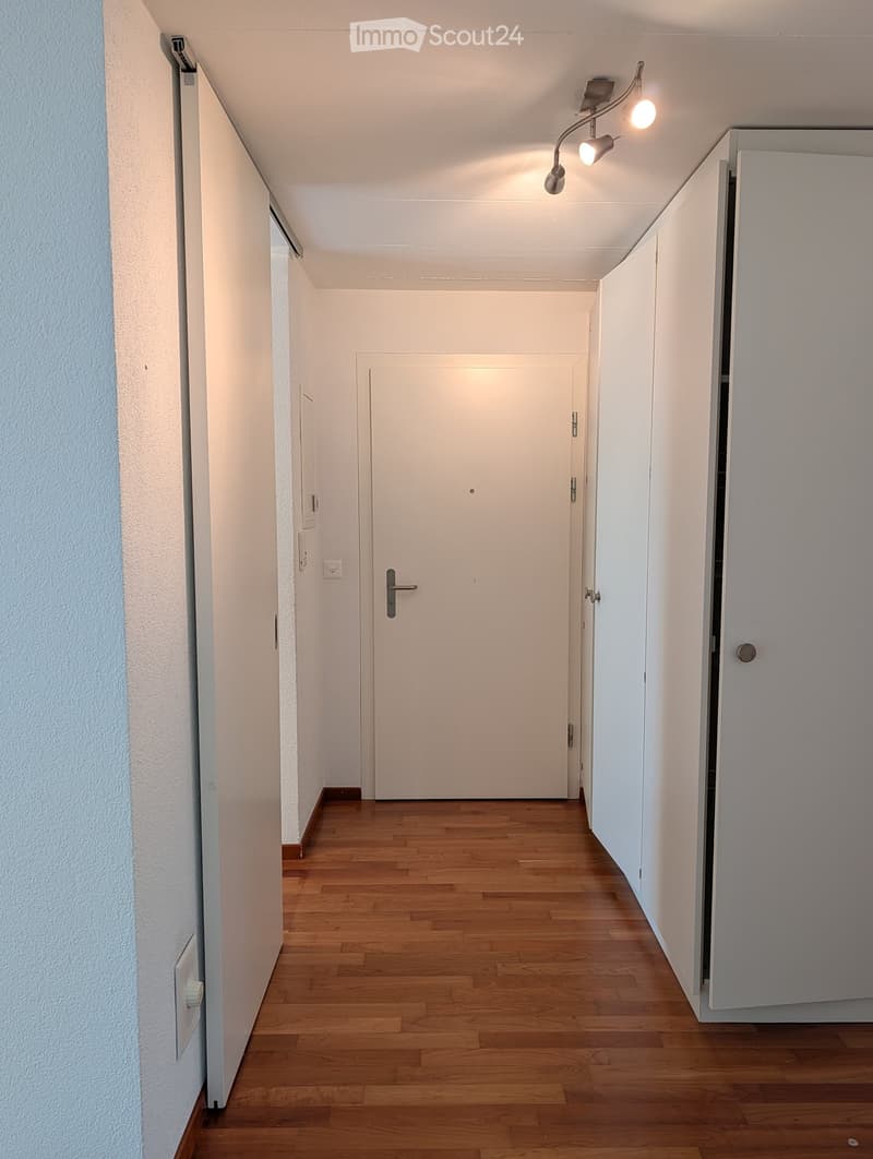 A clean, well-lit hallway with wooden flooring, white walls, and a closed door at the end.