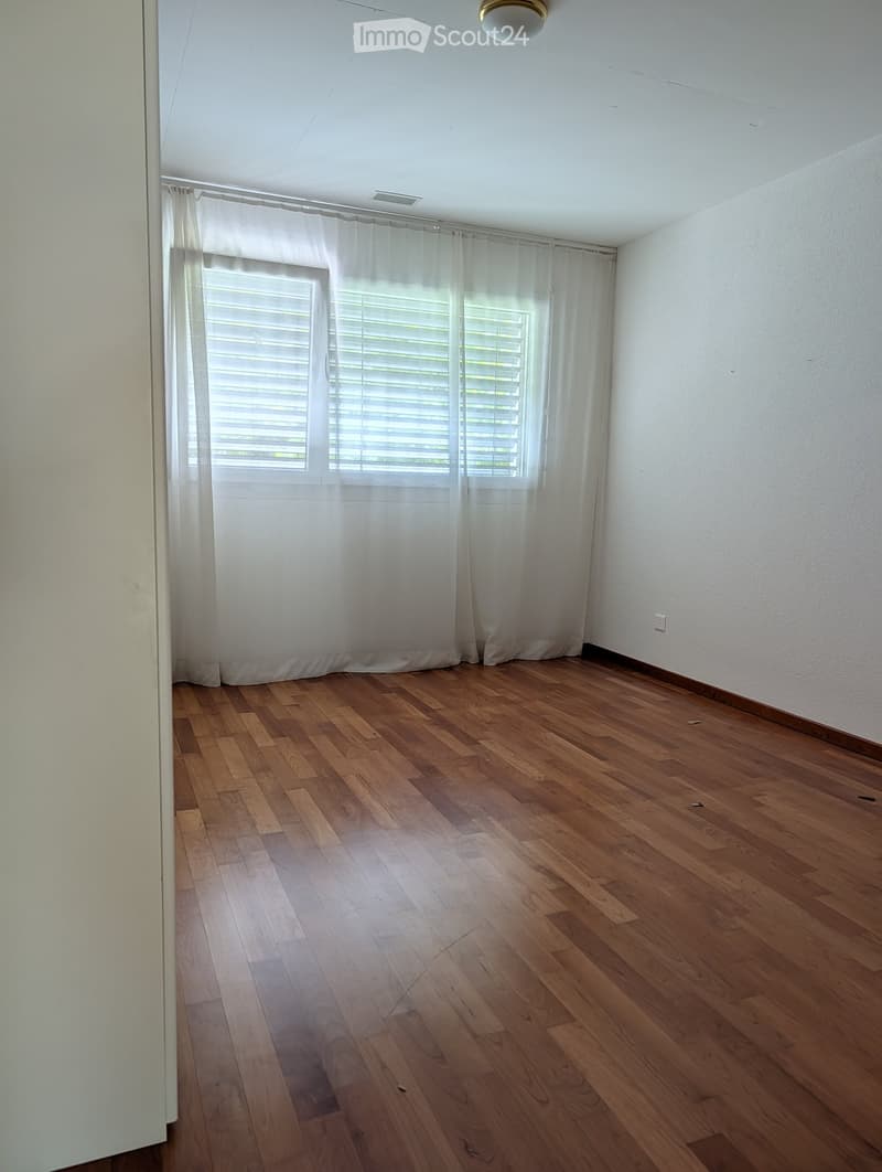 Empty room with wooden flooring, white walls, and large windows with blinds and curtains.