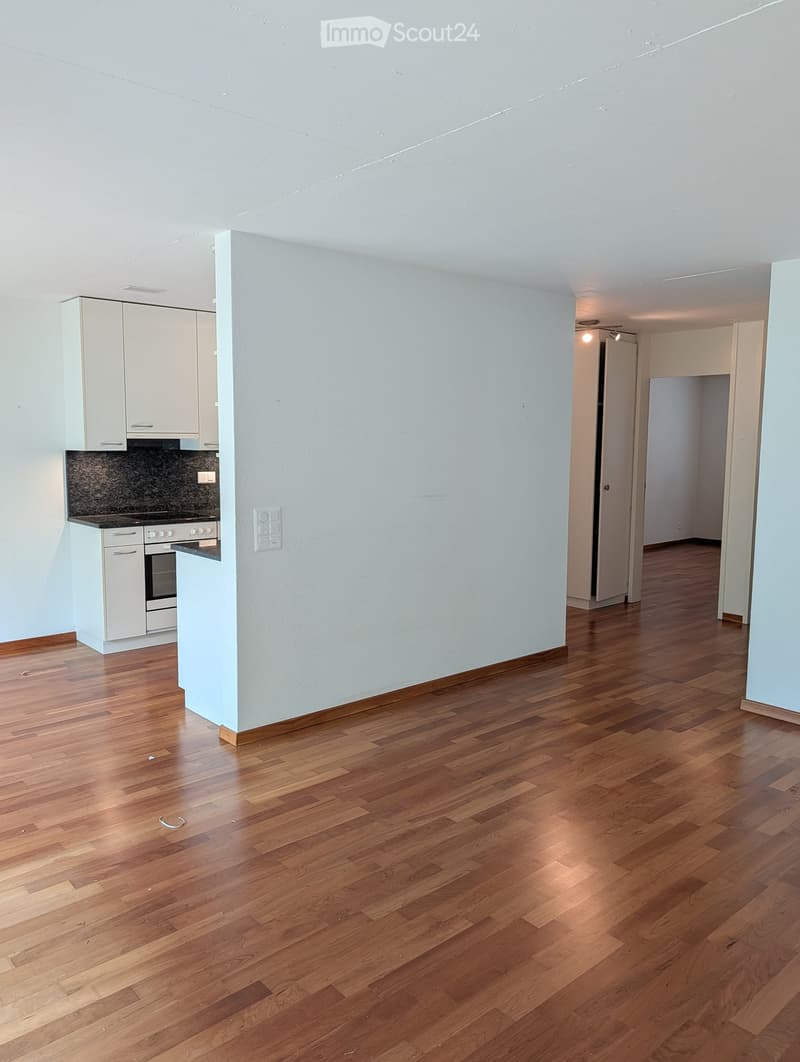 Empty room with light wooden flooring, white walls, and a small kitchen area with white cabinets and a countertop.