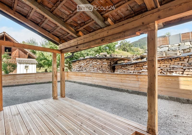 Wooden deck area with wooden beams and a stone woodpile, surrounded by wooden fencing and a gravel ground.