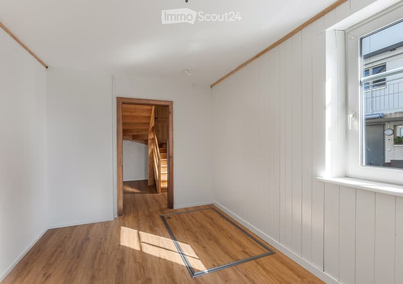 Bright hallway with wooden flooring, white walls, and a staircase leading upwards. Features a large window on the right side and a glass panel on the floor.