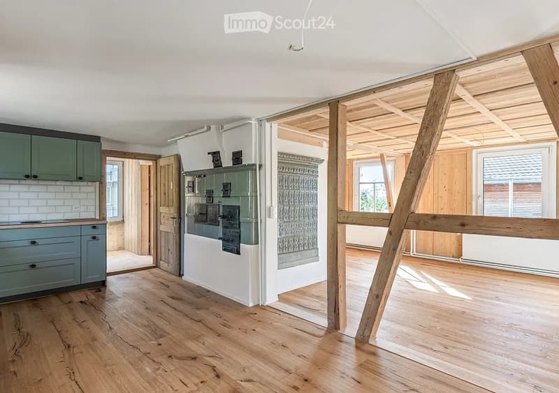 Empty room with exposed wooden beams and hardwood flooring, featuring a kitchen area with green cabinets and a stove, and a doorway leading to another room.
