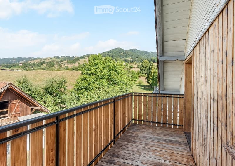 Wooden balcony with black railing, overlooking a scenic countryside with fields, trees, and distant hills.