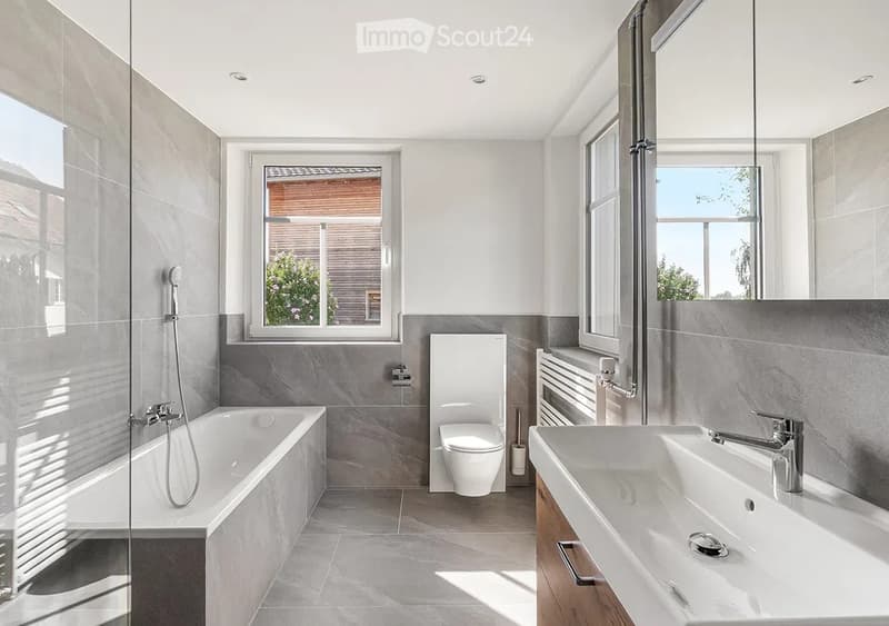 Modern bathroom with a bathtub, toilet, double sink, and large windows providing natural light. Features include a showerhead, radiator, and sleek grey tiles.