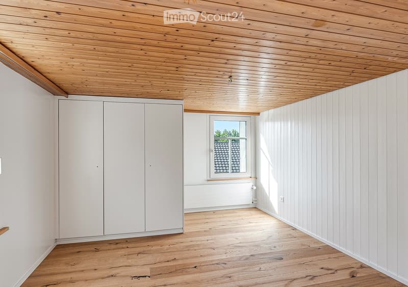 Empty room with wooden flooring, white paneled walls, and a large window showing a view of a roof. The ceiling is wooden with recessed lighting.