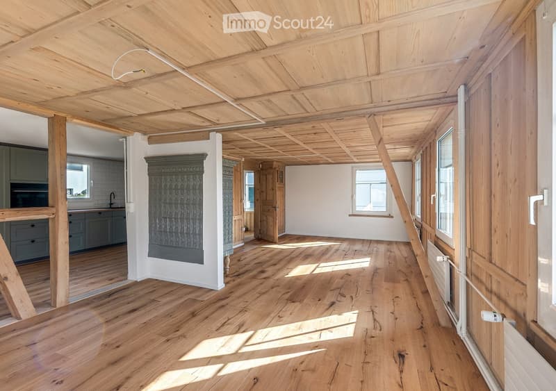 Bright, open room with exposed wooden beams and polished wooden flooring. Natural light streams in through multiple windows, creating a warm and airy atmosphere. The space features a fireplace with a decorative surround and a visible kitchen area with cab