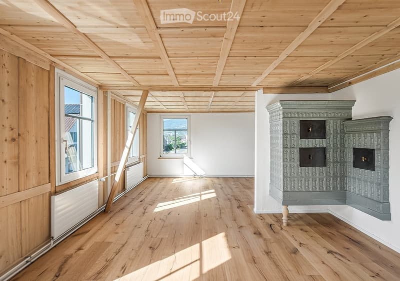 Empty room with wooden floor, exposed wooden ceiling beams, large windows, and a decorative fireplace.