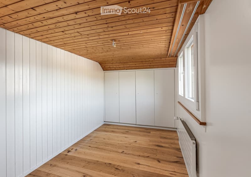 Empty room with light wooden flooring, white paneled walls, and a built-in white closet unit. The ceiling is exposed wooden beams, and there is a window with a wooden sill on one wall.