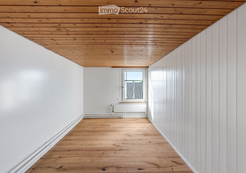 Empty hallway with wooden flooring, white walls, and a wooden ceiling. A single window at the end allows natural light to enter.