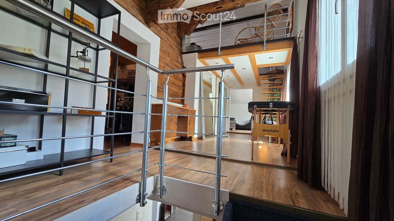 A modern, open-concept living space with wooden floors, exposed wooden beams, and a metal railing leading to an upper level. The room features built-in shelves, a wooden chair, a small table, and a curtained window area.