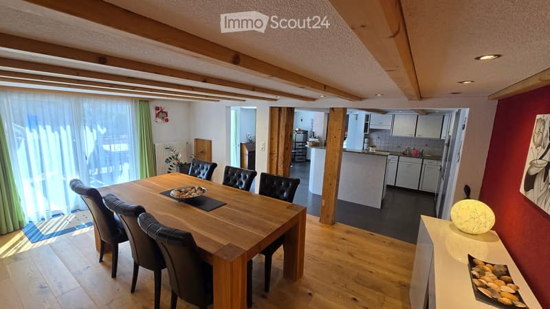 Bright dining area with wooden table and black leather chairs, adjacent modern kitchen with white cabinets, stainless steel appliances, and a small decorative lamp on a console table.