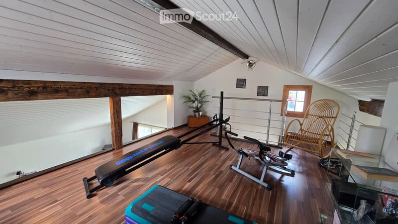 Well-lit room with wooden flooring, exercise bike, bench, wicker chair, potted plant, and a small table with books.