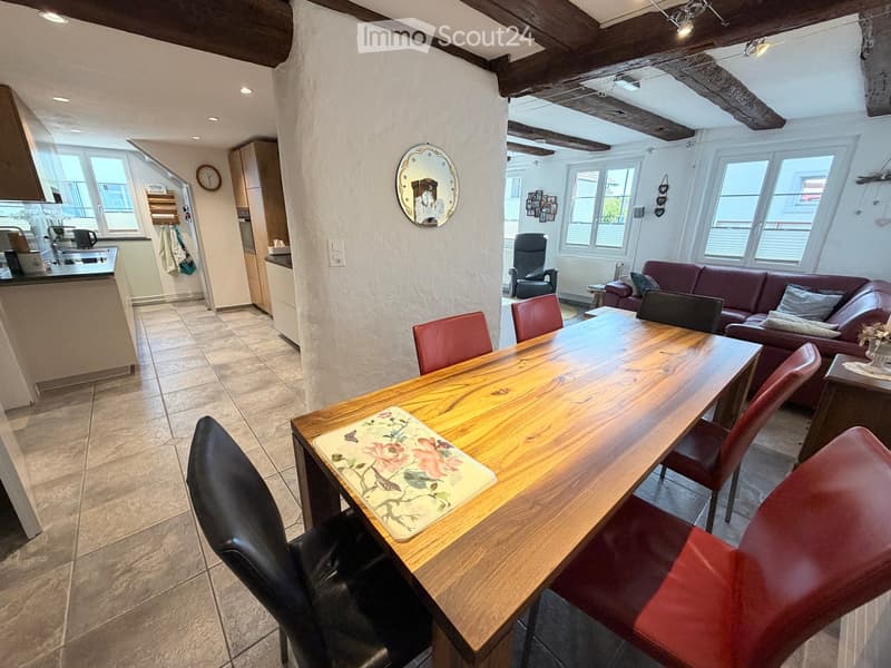 Open-plan living and dining area with exposed wooden beams, a large wooden dining table with red and black chairs, a red leather sofa, a kitchen area with a counter, and a decorative mirror on the wall.