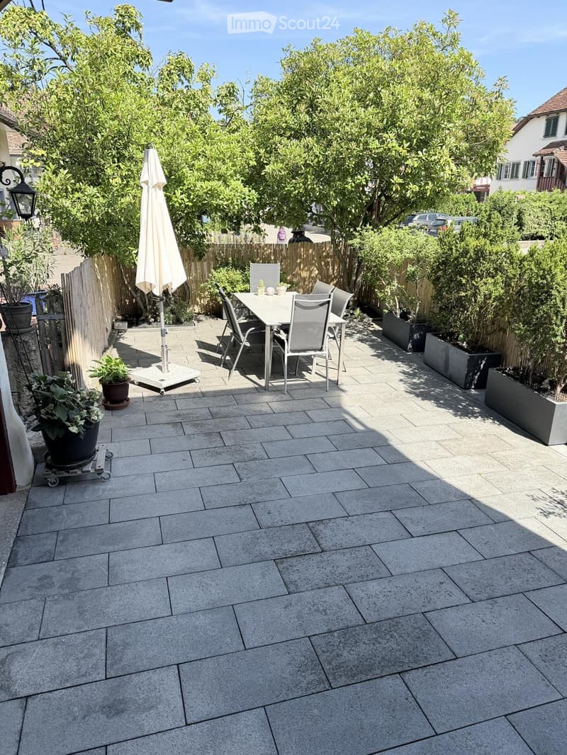 Outdoor patio with stone tiles, a white table and chairs, an umbrella, and several potted plants.