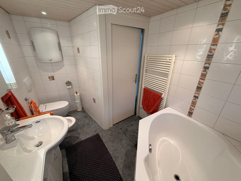 Modern bathroom with white tiles, a bathtub, a sink with a faucet, a toilet, a towel rack with towels, and a mirror above the sink.