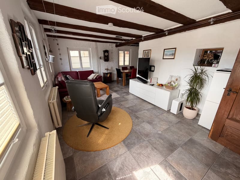 Living room with exposed wooden beams, tiled floor, sofa, armchair, coffee table, TV, dining table, and potted plant.