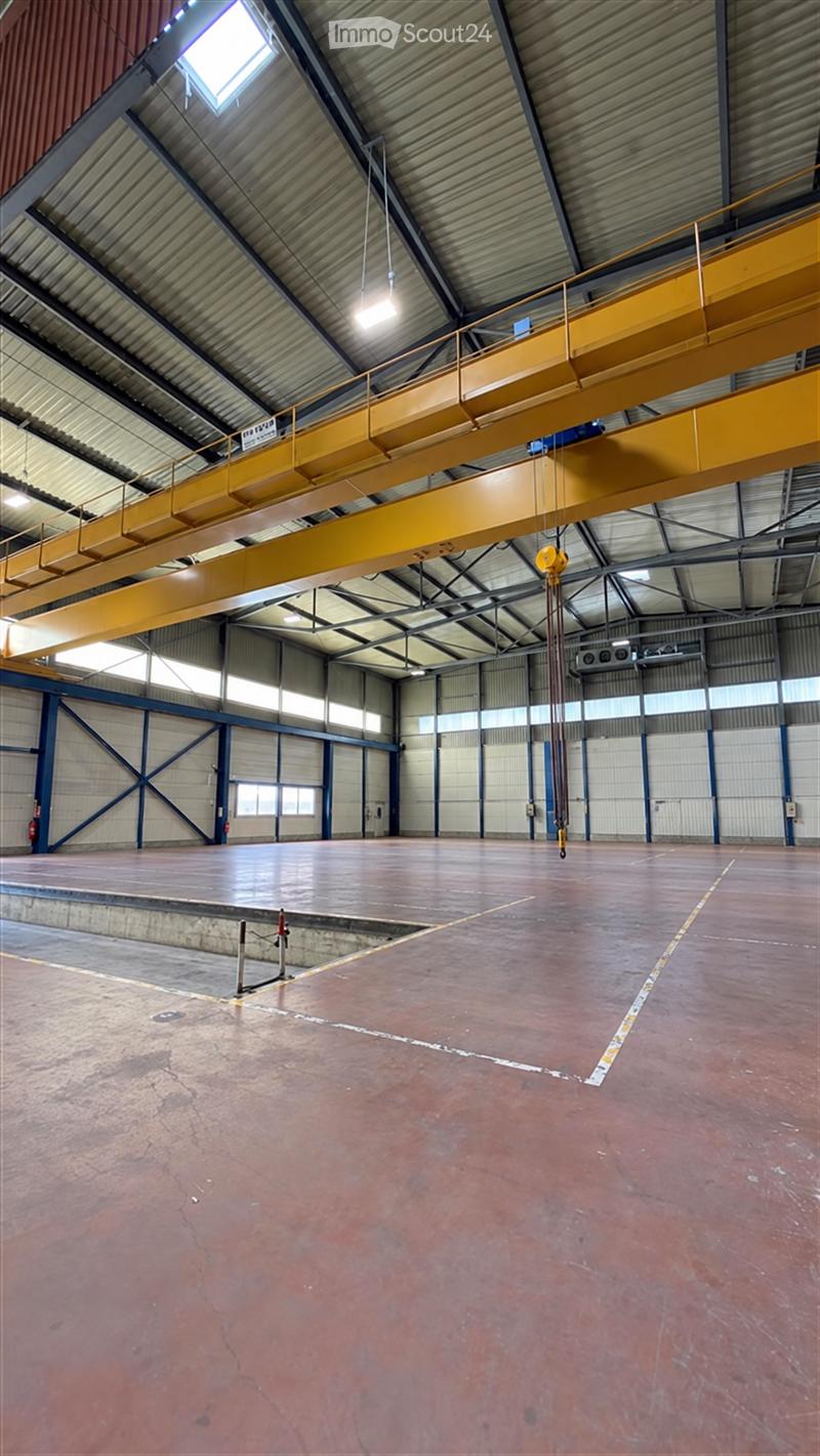 Large industrial-style warehouse space with a high, metal-truss ceiling, exposed beams, and a yellow overhead crane system. The floor is concrete, and the walls are metal with large windows. The space is empty, with no furniture or equipment.