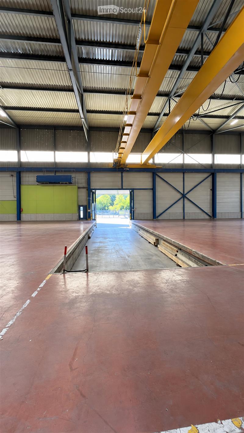 Large industrial-style warehouse space with a high ceiling, metal framework, and a red concrete floor. The area is empty, with no furniture or equipment visible.