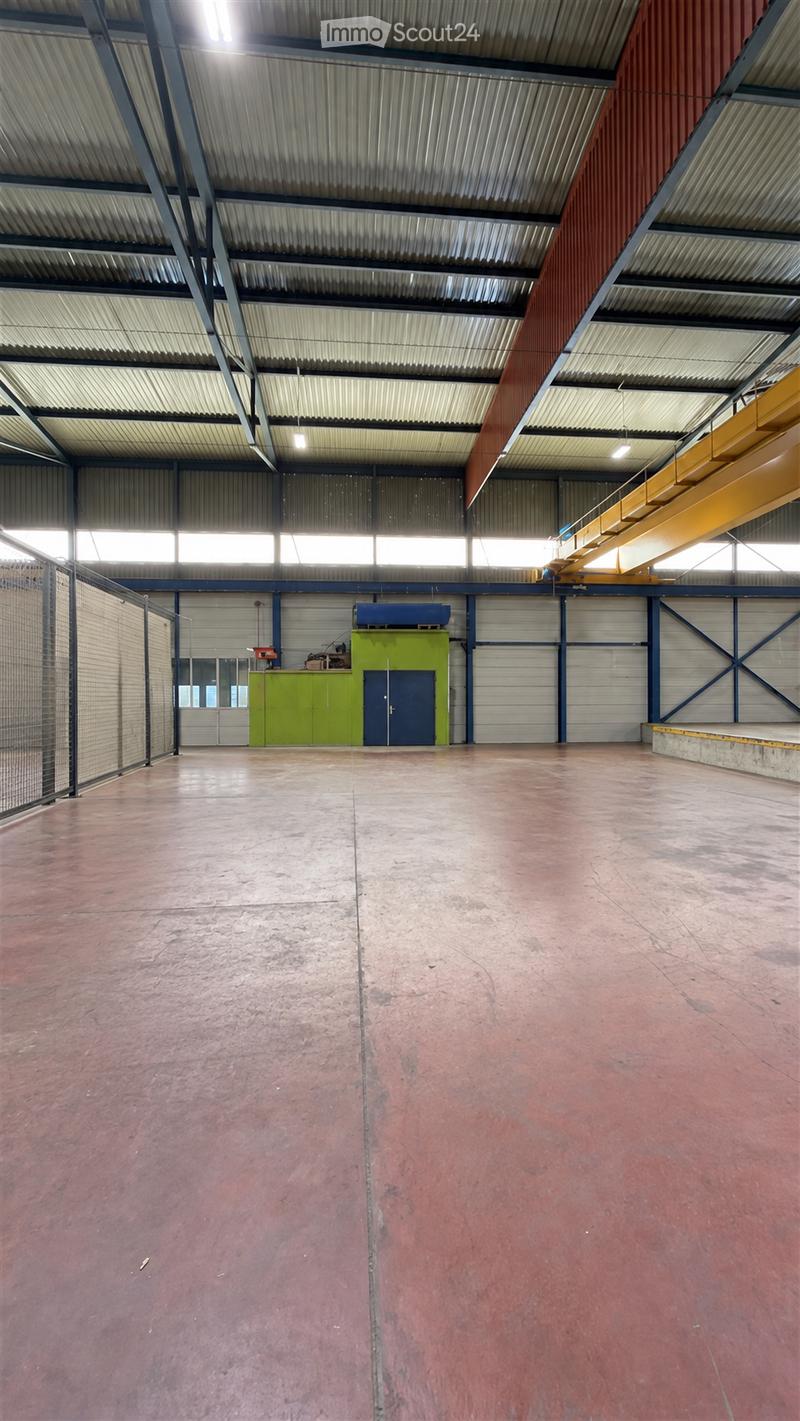 Large, empty industrial-style warehouse with high ceilings, metal walls, and a concrete floor. Features include large windows, a green utility cabinet, and a blue door.