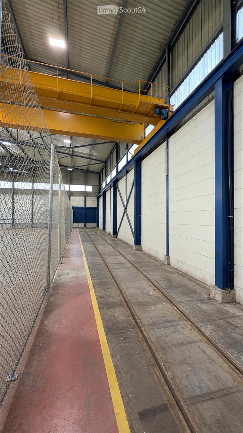 Industrial-style corridor with metal railings, overhead crane, and large windows. The floor is concrete with a red carpeted section on one side.