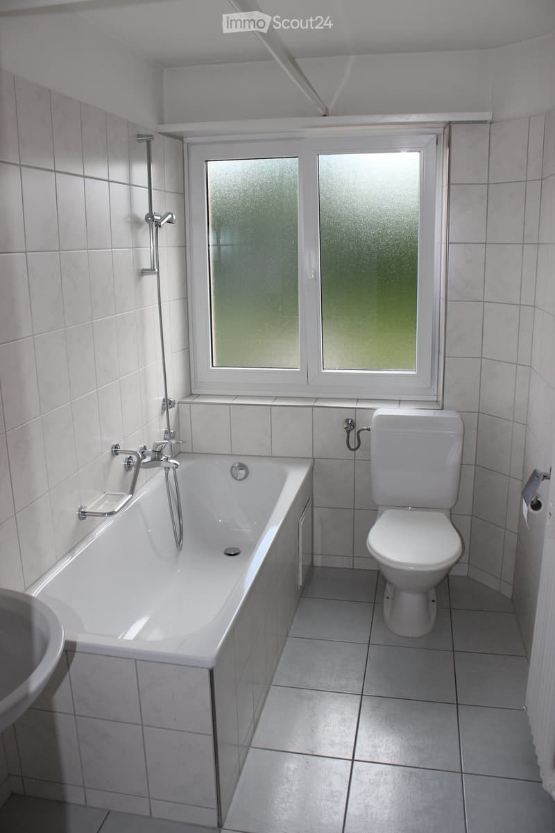 Modern bathroom with white tiled walls and floor, featuring a bathtub, toilet, and a window with frosted glass panes.