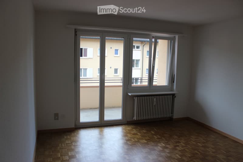 Empty room with wooden flooring, large sliding glass doors leading to a balcony, radiator, and visible electrical outlets.