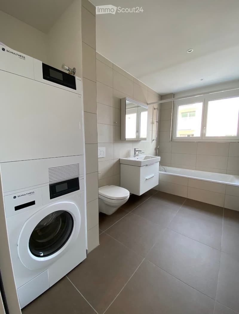 Modern bathroom with a washing machine, toilet, sink, bathtub, and mirror. The space features tiled walls and a window providing natural light.