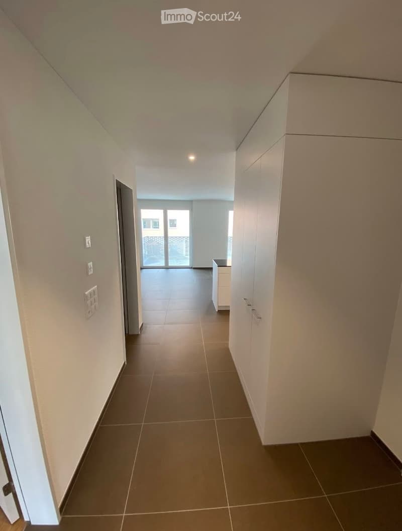 A clean, modern hallway with tiled flooring, white walls, and built-in storage cabinets on one side. The hallway leads to a room with large glass doors and a view of the outside.